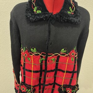 Könnte beinhalten: Schwarze Strickjacke mit rotem Karomuster auf der Vorderseite. Der Pullover hat einen schwarzen Kragen mit floralen und perlenbesetzten Details. Florale Verzierungen befinden sich auch an den Manschetten und Taschen. Die Strickjacke ist mit Knöpfen versehen.