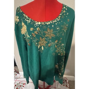 Könnte beinhalten: Ein grüner Weihnachtspullover mit goldenen Pailletten und Perlenverzierungen. Der Pullover zeigt Rentier-, Poinsettien- und Schneeflocken-Designs. Der Halsausschnitt ist mit goldenen Pailletten verziert.