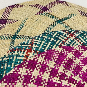 Puede incluir: Una pieza decorativa circular tejida con una base beige natural y patrones geométricos en verde azulado, morado y rojo. Los patrones se crean tejiendo hebras de colores en la base de fibra natural.
