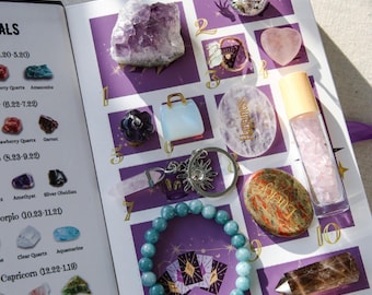 Op de afbeelding: Een open boek met een paarse en witte lay-out, met verschillende kristallen, een armband en een kleine boomsculptuur. De kristallen omvatten amethist, rozenkwarts en bergkristal. Een armband met blauwe kralen is ook zichtbaar.