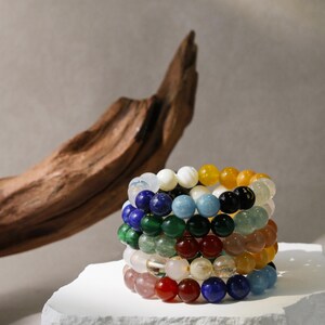 May include: A stack of beaded bracelets in various colors, including red, blue, green, and yellow. The bracelets are made of polished gemstones and are stacked on a white surface. A piece of driftwood is in the background.