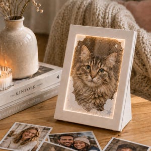 Pode incluir: Uma moldura de foto branca e iluminada exibindo um retrato de gato, com um design de nuvens e estrelas. Várias fotografias impressas de pessoas e um gato estão espalhadas na superfície de madeira. Um vaso com flores secas e uma vela também são visíveis.