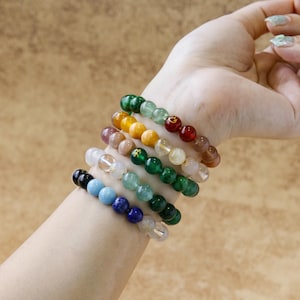 Puede incluir: Una colección de pulseras de cuentas de varios colores, incluyendo verde, amarillo, rojo y azul. Las pulseras están hechas de cuentas redondas y están apiladas en una muñeca. El fondo es de color marrón claro.