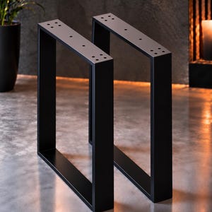 Könnte beinhalten: Ein Paar Tischbeine aus mattschwarzem Metall mit rechteckigem Rahmendesign. Jedes Bein verfügt über eine flache Montageplatte an der Oberseite mit vorgebohrten Löchern zur Befestigung. Die Struktur besteht aus vertikalen und horizontalen Stangen.