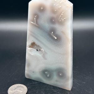 May include: A polished, rectangular agate crystal obelisk with a flat top. The stone features a mix of white, gray, and teal colors with dark gray circular patterns. A quarter coin is next to the obelisk for size comparison.