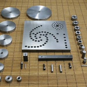 May include: A set of metal tools for crafting and jewelry making. The set includes a variety of round metal discs, a square metal plate with a spiral pattern of holes, a metal bar, and a collection of metal cylinders.