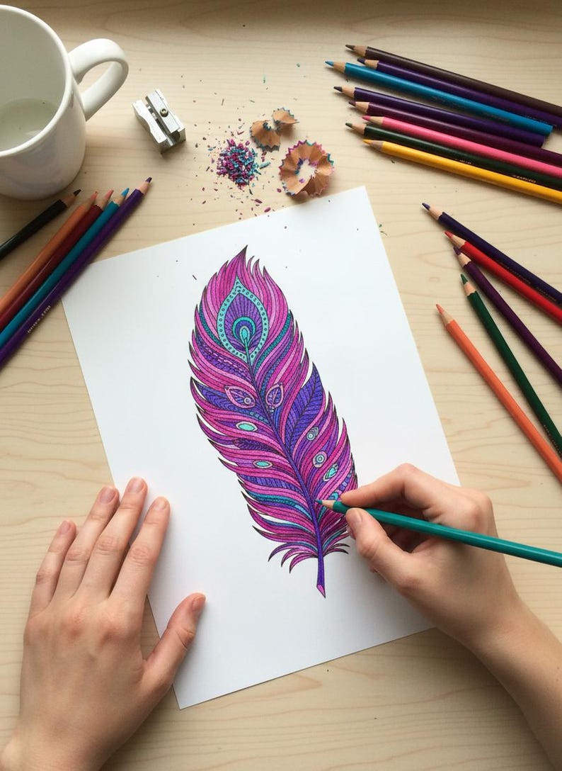 A lifestyle preview of a person's hands coloring a detailed single peacock feather with a teal pencil. This 60-page collection showcases high-quality bohemian line art featuring vibrant magenta, purple, and turquoise patterns. The scene is set on a light wooden desk with art supplies, perfect for promoting adult relaxation, mindful meditation, and printable Zen activities.