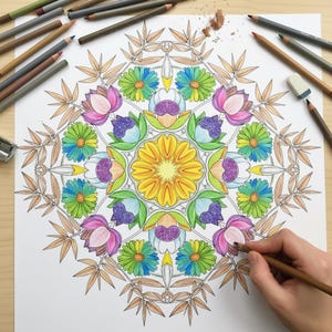 A person's hand coloring an intricate floral mandala featuring a vibrant yellow center, green-and-blue daisies, and pink lotus flowers. This lifestyle preview, showing colored pencils and shavings on a wooden desk, highlights the relaxing and creative experience of our 100-page Floral Mandala collection, perfect for adult relaxation, mindfulness, and stress relief.