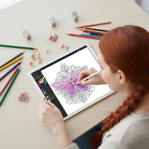 May include: A digital tablet displaying a butterfly coloring page with pink and purple hues. Colored pencils, pencil sharpeners, and pencil shavings are scattered on the light-colored wooden surface. A stylus is being used to color the butterfly.