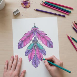 A lifestyle preview of a person's hands coloring a symmetrical boho feather arrangement with a teal colored pencil. This 60-page collection features intricate pink, purple, and turquoise patterns on a wooden desk setting with art supplies, showcasing high-quality line art designed for adult relaxation, meditation, and printable creative activities.