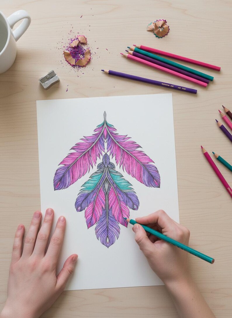 A lifestyle preview of a person's hands coloring a symmetrical boho feather arrangement with a teal colored pencil. This 60-page collection features intricate pink, purple, and turquoise patterns on a wooden desk setting with art supplies, showcasing high-quality line art designed for adult relaxation, meditation, and printable creative activities.