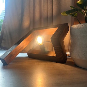 Könnte beinhalten: Eine moderne Tischlampe aus Holz mit geometrischem Design. Die Lampe hat eine warme, leuchtende Glühbirne in einem dunkelbraunen, eckigen Holzrahmen. Die Lampe steht auf einer hellen Oberfläche.