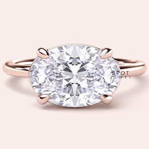 May include: Rose gold engagement ring featuring an oval-cut diamond. The diamond is held in place by four prongs. The ring band is a simple, polished design. The text "SPOT JEWEL" is visible on the right side of the ring.