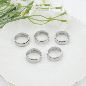 May include: Five silver-toned metal rings with smooth, polished surfaces. Each ring features a unique name engraved in black lettering on the outer band. The names visible on the rings are Sylus, Rafayel, Zayne, Xavier, and Caleb. The rings are circular.