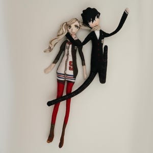 May include: Two handmade fabric dolls. One doll has blonde hair, a white dress with a red and blue stripe design, and red tights. The other doll is wearing a black suit and has black hair. The dolls are embracing.