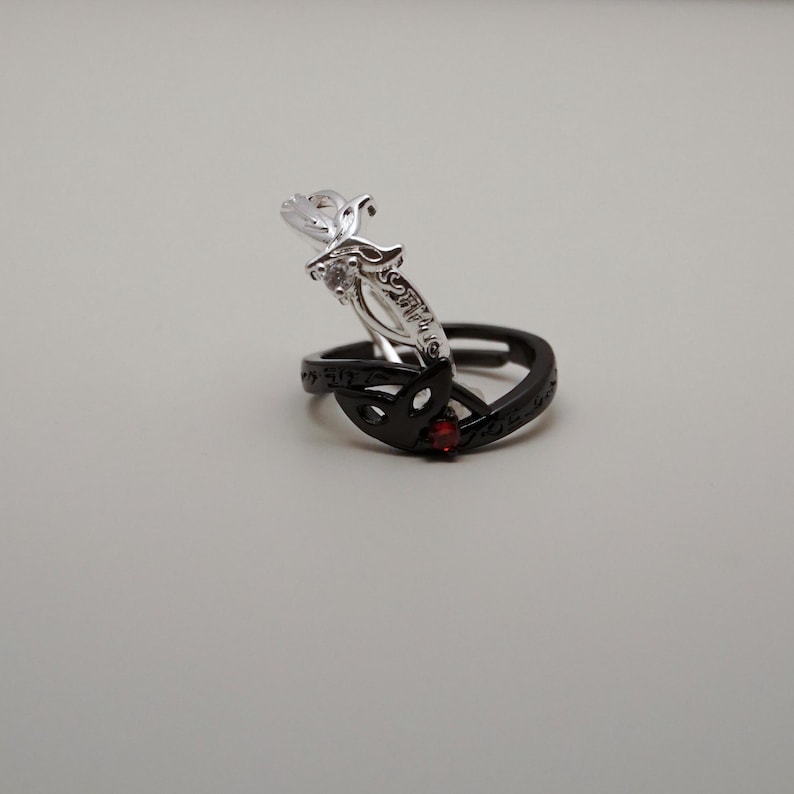 K&ouml;nnte beinhalten: Gezeigt werden zwei Ringe, einer silberfarben und einer schwarz. Der silberfarbene Ring hat ein dekoratives Design mit einem klaren Stein. Der schwarze Ring hat ein ausgeschnittenes Design und einen kleinen roten Stein. Die Ringe sind vor neutralem Hintergrund.