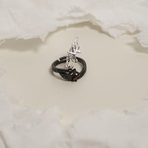 K&ouml;nnte beinhalten: Zwei Ringe, einer schwarz und einer silberfarben, werden gezeigt. Der schwarze Ring hat ein Blattdesign mit einem kleinen roten Edelstein. Der silberne Ring hat eine dekorative Figur. Beide Ringe liegen auf einer wei&szlig;en Oberfl&auml;che.