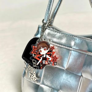 Peut inclure: Un porte-clés en acrylique transparent représentant un personnage d'anime stylisé aux cheveux bruns et aux yeux rouges, vêtu d'une tenue noire. Le design inclut des étoiles rouges et le texte "MAKOTO" en gras. Il est fixé à un fermoir en métal argenté.