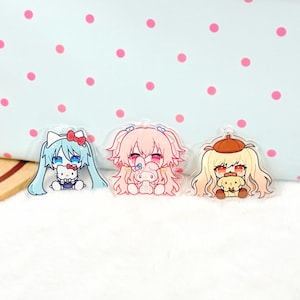 May include: Three clear acrylic charms featuring chibi-style characters. The left charm shows a blue-haired figure with a white cat, the centre charm depicts a pink-haired figure with a pink rabbit, and the right charm shows a blonde figure with a dog.