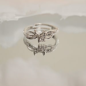 K&ouml;nnte beinhalten: Ein silberfarbener Ring mit floralem Design, besetzt mit kleinen, klaren Edelsteinen. Der verstellbare Ring spiegelt sich auf einer spiegelnden Oberfl&auml;che wider und zeigt seine komplizierten Details und seine Handwerkskunst. Das Ringdesign umfasst ein zentrales Blumenelement.