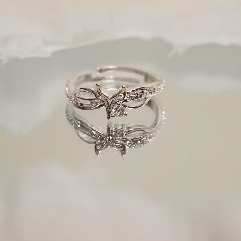 K&ouml;nnte beinhalten: Ein silberfarbener Ring mit floralem Design, besetzt mit kleinen, klaren Edelsteinen. Der verstellbare Ring spiegelt sich auf einer spiegelnden Oberfl&auml;che wider und zeigt seine komplizierten Details und seine Handwerkskunst. Das Ringdesign umfasst ein zentrales Blumenelement.