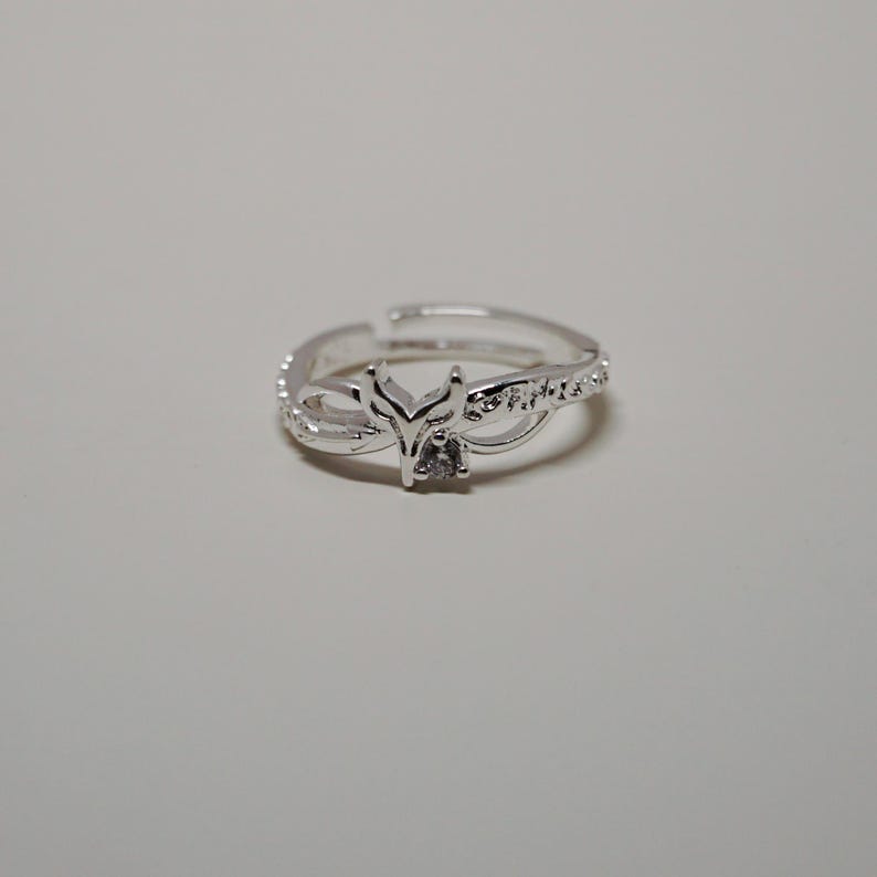 K&ouml;nnte beinhalten: Ein silberfarbener Ring mit einem Fuchskopf-Design. Der Ring hat einen kleinen, quadratisch geschliffenen, klaren Stein in der Mitte des Fuchskopfes. Das Band ist mit kleinen, klaren Steinen und dekorativen Wirbeln verziert.