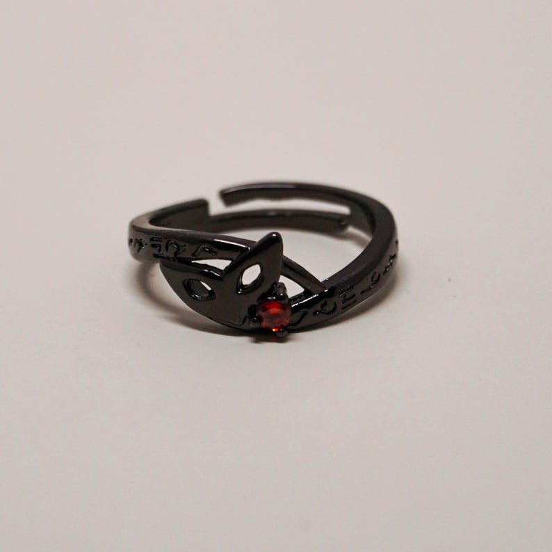 K&ouml;nnte beinhalten: Verstellbarer schwarzer Ring mit einem Fuchskopf-Design und einem kleinen roten Edelstein. Der Ring hat ge&auml;tzte Details und ist vor einem wei&szlig;en Hintergrund platziert. Ein einzigartiges Schmuckst&uuml;ck.