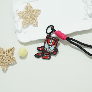 May include: A red and black cartoon character keyring with a blue and white face design. The keyring is attached to a black lanyard with a pink accent. Woven stars and a yellow pom-pom are also in the image.