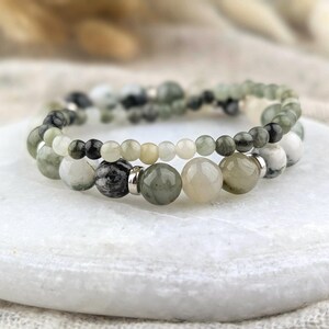May include: A double-strand bracelet featuring smooth, round beads in shades of sage green, charcoal gray, cream, and white. The design incorporates smaller beads on the top strand and larger beads on the bottom strand, accented with small silver-toned metal spacers.