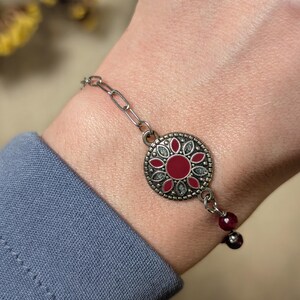 Peut inclure: Un bracelet à chaîne argentée doté d'un pendentif circulaire avec un centre en émail rouge entouré d'un motif floral de pétales rouges et d'accents argentés texturés. Le bracelet comprend deux petites perles sphériques rouge foncé.