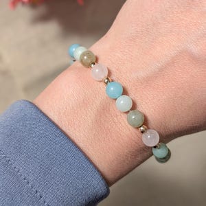 May include: A beaded bracelet featuring alternating round polished stones in shades of light blue, pale pink, and muted green, interspersed with small metallic gold-toned spherical spacers, strung on a clear elastic cord worn around the wrist.