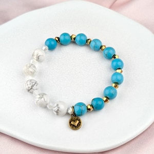 May include: A beaded bracelet featuring turquoise and white marble-like beads, interspersed with gold-colored spacers. A small, gold-toned charm with a heart design hangs from the bracelet. The bracelet is displayed on a white plate.