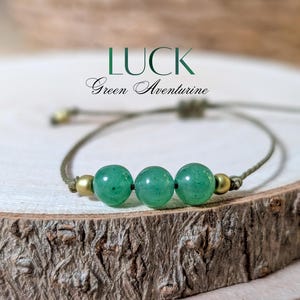 May include: A bracelet featuring three round green aventurine beads flanked by two small metallic gold-toned beads on a thin olive green cord. The text LUCK and Green Aventurine is printed above the bracelet in dark green and black script.