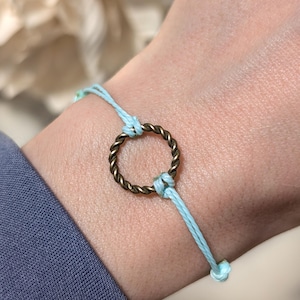 May include: A light blue cord bracelet featuring a central circular charm made of twisted antique brass metal. The cord is tied securely around the metal ring, creating a simple and clean design that sits flat against the wrist area.