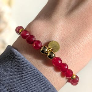 Könnte beinhalten: Ein Perlenarmband mit facettierten, dunkelroten, durchscheinenden Perlen, die mit polierten, goldfarbenen Kugeln durchsetzt sind. Ein kleiner, flacher, kreisförmiger goldfarbener Anhänger hängt in der Mitte des am Handgelenk getragenen Armbands.