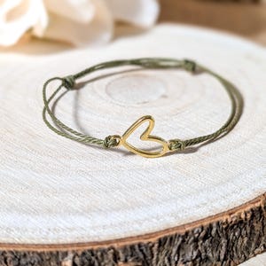 May include: A bracelet featuring a thin, olive green cord and a central gold-toned metal heart charm. The cord is tied with adjustable knots on either side of the heart, creating a simple, continuous loop design with a smooth, polished finish.
