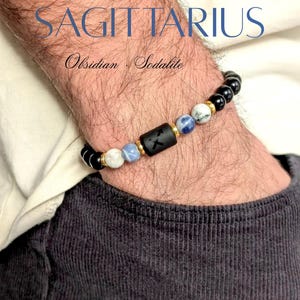 Op de afbeelding: Een kralenarmband met zwarte obsidiaan, blauw-witte sodaliet en goudkleurige tussenstukjes. Een centrale matzwarte cilindrische kraal toont het sterrenbeeld Boogschutter. De tekst SAGITTARIUS en Obsidian · Sodalite is zichtbaar.