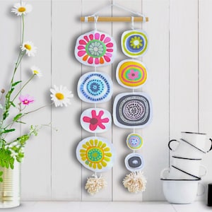 Könnte beinhalten: Bunte Wanddekoration mit abstrakten Blumenmustern in verschiedenen Formen und Farben. Die Dekoration wird an einem Holzstab aufgehängt. Eine Vase mit Blumen und ein Stapel weißer Tassen mit schwarzen Rändern und Griffen sind ebenfalls zu sehen.