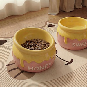 Peut inclure: Deux gamelles pour animaux de compagnie en c&eacute;ramique avec un design de pot de miel. Les bols sont jaunes et roses, avec le mot "HONEY" sur l'un et "SWEE" sur l'autre. L'un des bols est rempli de nourriture brune pour animaux.