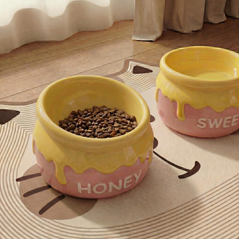 Peut inclure: Deux gamelles pour animaux de compagnie en c&eacute;ramique avec un design de pot de miel. Les bols sont jaunes et roses, avec le mot "HONEY" sur l'un et "SWEE" sur l'autre. L'un des bols est rempli de nourriture brune pour animaux.