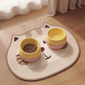 Peut inclure: Un tapis de nourriture pour animaux de compagnie en forme de chat avec deux bols jaunes et roses. Un bol contient de la nourriture pour animaux, l'autre est vide. Le tapis a un visage de chat et les mots "HONEY" et "SWEET" sont sur les bols.