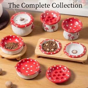 May include: A collection of ceramic pet bowls and feeders featuring red and white mushroom designs with polka dots. The set includes various bowl shapes, some with raised textures, and a rectangular wooden stand holding two bowls filled with kibble.