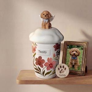 May include: A white ceramic pet urn with a floral design and the name "Teddy". A small dog figurine with angel wings sits on top. A framed photo and paw print keepsake are also present.