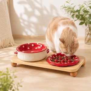 May include: A ceramic pet feeding station featuring a raised wooden platform with four small ball-shaped feet. It holds a round white bowl with a red and white polka dot interior and a smaller red and white polka dot slow feeder bowl with bumps.