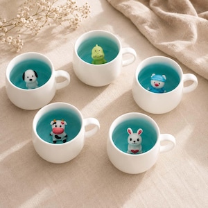 Pode incluir: Cinco canecas de cerâmica branca com interior turquesa brilhante, cada uma com uma pequena figura de animal tridimensional no fundo. As figuras incluem um cão, um dinossauro verde, um porco com chapéu azul, uma vaca e um coelho branco segurando um coração vermelho.