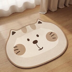 Peut inclure: Un tapis de sol beige et marron en forme de chat. Le tapis pr&eacute;sente un visage de chat souriant avec des accents marron pour les yeux, le nez et les joues. Le tapis a une bordure ray&eacute;e et est pos&eacute; sur un sol en bois.