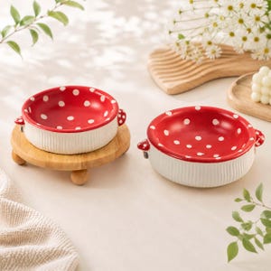 May include: Two round ceramic bowls featuring a vibrant red interior with white polka dots and a textured white exterior. Each bowl has small side handles and a shallow design. One bowl rests on a small, round wooden stand with three feet.
