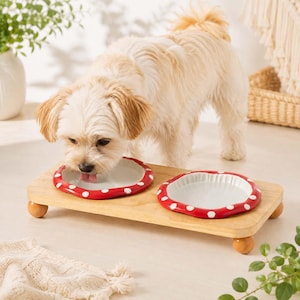 May include: A light-colored dog eating from a raised wooden feeder featuring two white ceramic bowls with red rims and white polka dots. The rectangular wooden stand is supported by four small, rounded wooden feet, providing a stable base.