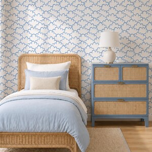 May include: A bedroom scene featuring a light blue bedspread and pillows on a wicker bed frame. A blue dresser with woven panels and a white lamp sit beside the bed. The wallpaper has a repeating blue cloud pattern, and a beige rug covers the floor.