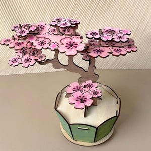 May include: A wooden bonsai tree ornament featuring pink cherry blossom flowers. The tree is set in a light green and beige pot. The trunk and branches are brown.